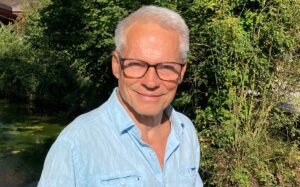 Ein Mann mit blauen Hemd steht vor einem grünen Hintergrund und lächelt|