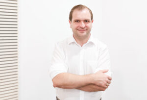 Kardiologe Dr. Arnulf Holzknecht im Portrait vor einer weißen Wand mit weißem Hemd und die Hände vor dem Körper verschrenkt.